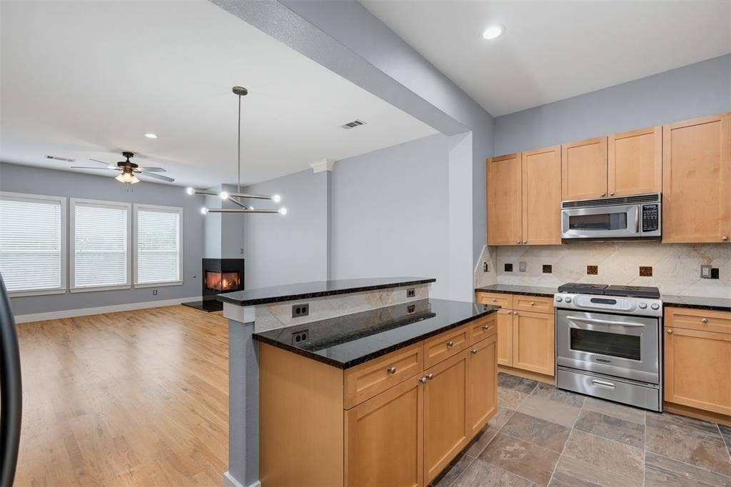 4830 Cedar Springs Road, #30, Dallas, TX 75219 Kitchen and living area featuring a two-tiered island with a black countertop, natural wood-finish cabinetry, stainless steel appliances, and a tiled backsplash