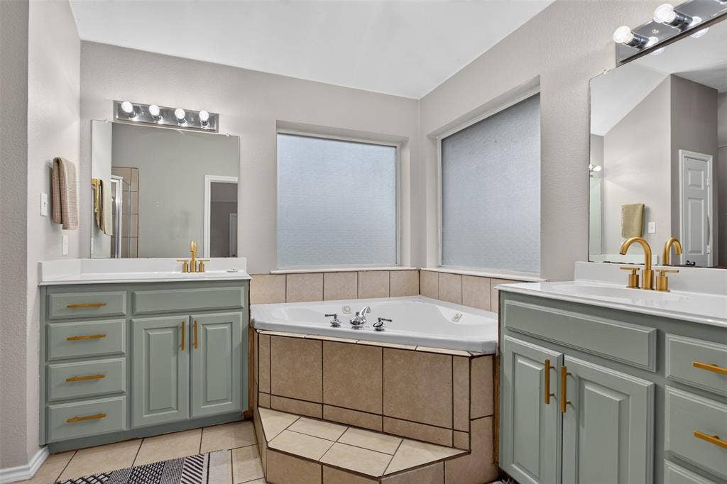 Corner soaking tub with tiled surround, flanked by two vanities featuring light green cabinetry, white countertops, and gold-toned fixtures