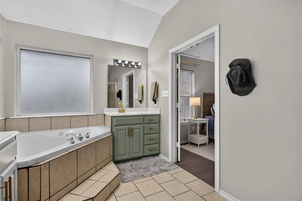 Soaking tub with tiled surround, light-toned tile flooring, dual-basin vanity with a light green cabinet finish and gold-toned hardware, and a vaulted ceiling