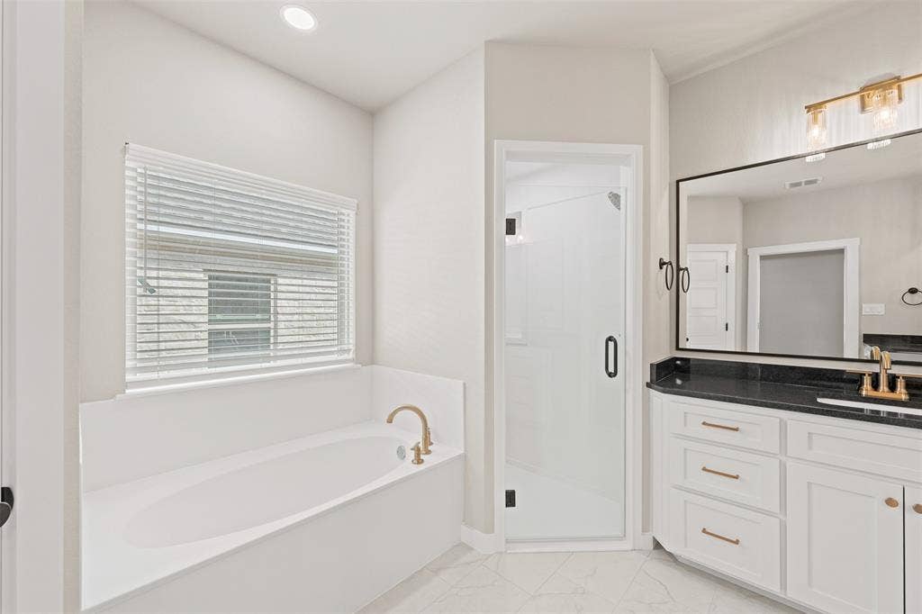 9824 Durango Trail, Waco, TX 76712 Bathroom featuring a built-in soaking tub with brass fixtures, a walk-in shower with a frameless glass door, a double vanity with a dark countertop and gold-toned hardware, and a large wall-mounted mirror