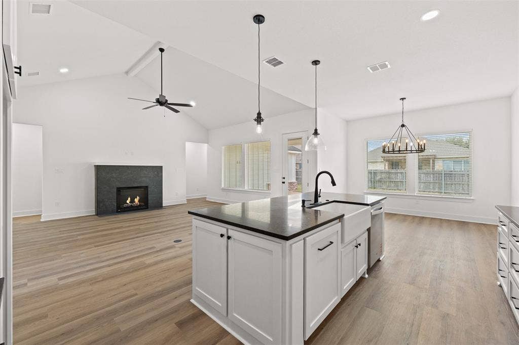 9820 Durango Trail, Waco, TX 76712 Open-concept interior featuring light wood-finish flooring, a kitchen island with a farmhouse sink and black countertop, a fireplace with a dark stone surround, vaulted ceilings, and multiple windows for natural light