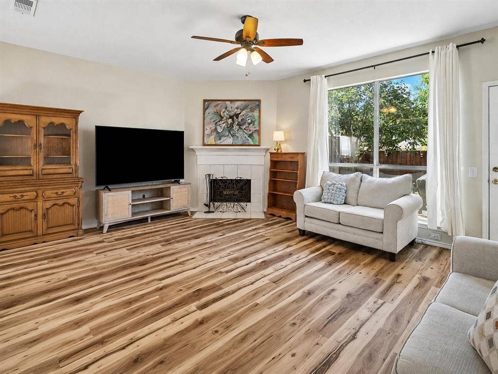 11379 Lockshire Drive, Frisco, TX 75035 Spacious living area featuring wood-finish flooring, a ceiling fan, a fireplace with a white mantel, and large windows