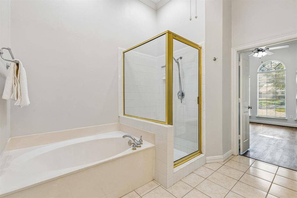 6852 High Field Trail, Plano, TX 75023 Bathroom featuring a built-in soaking tub, a stand-up shower with a glass enclosure and gold-toned frame, and light-toned tile flooring