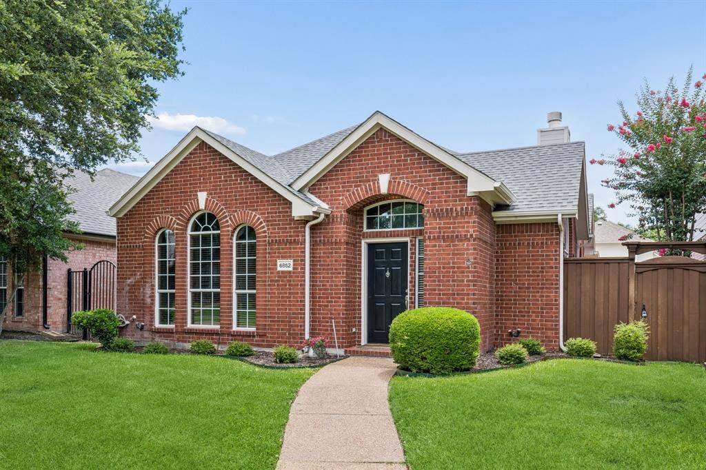 6852 High Field Trail, Plano, TX 75023 Red brick facade featuring a dark entry door with transom window, arched windows, a concrete pathway, a shingle roof, and a wood privacy fence