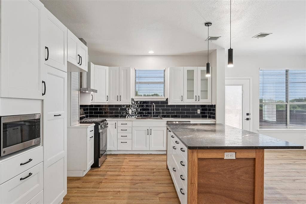 White cabinetry, black subway tile backsplash, stainless steel appliances, wood-finish flooring, and a kitchen island with pendant lighting