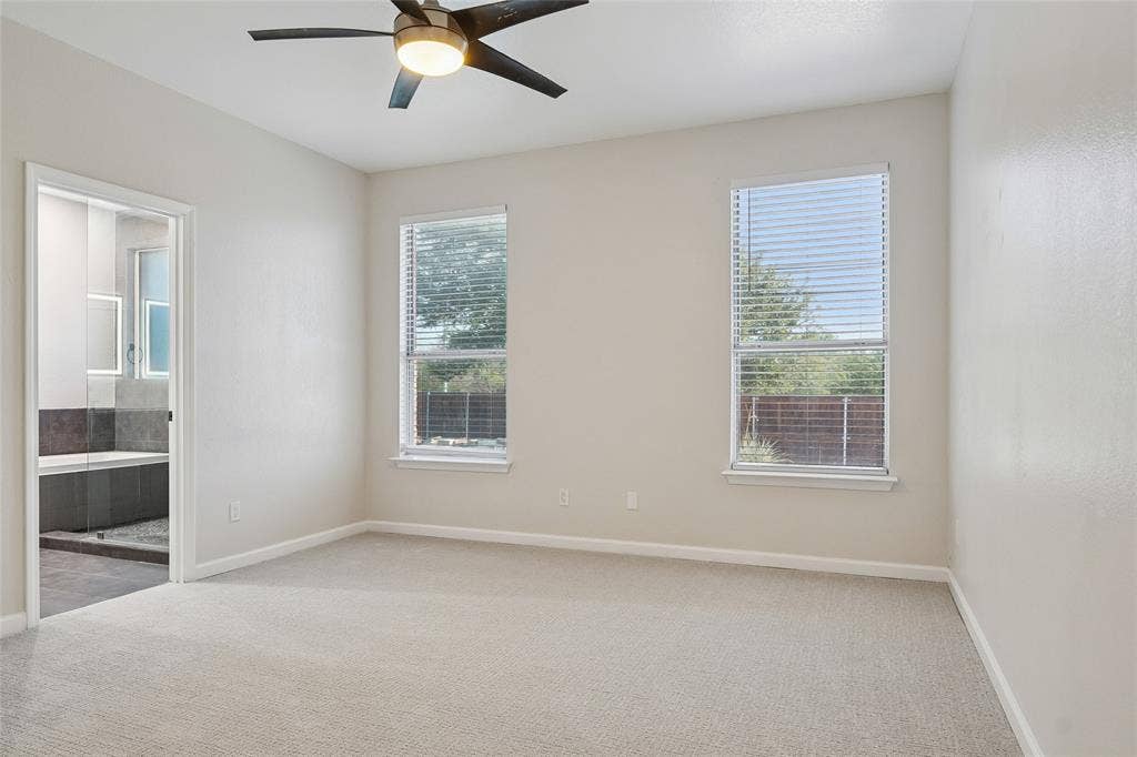 Carpeted flooring, two windows with blinds, modern ceiling fan with integrated lighting, and an attached full bathroom with a tiled tub
