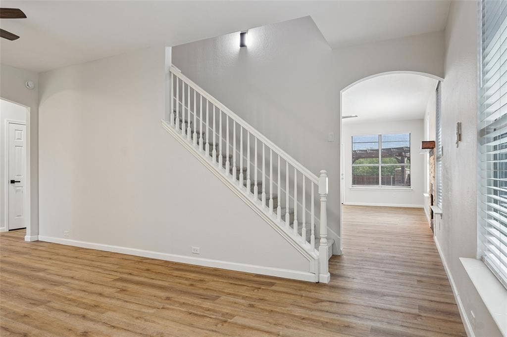 Wood-finish flooring, white painted walls, white spindle staircase, arched doorway, and a window with blinds