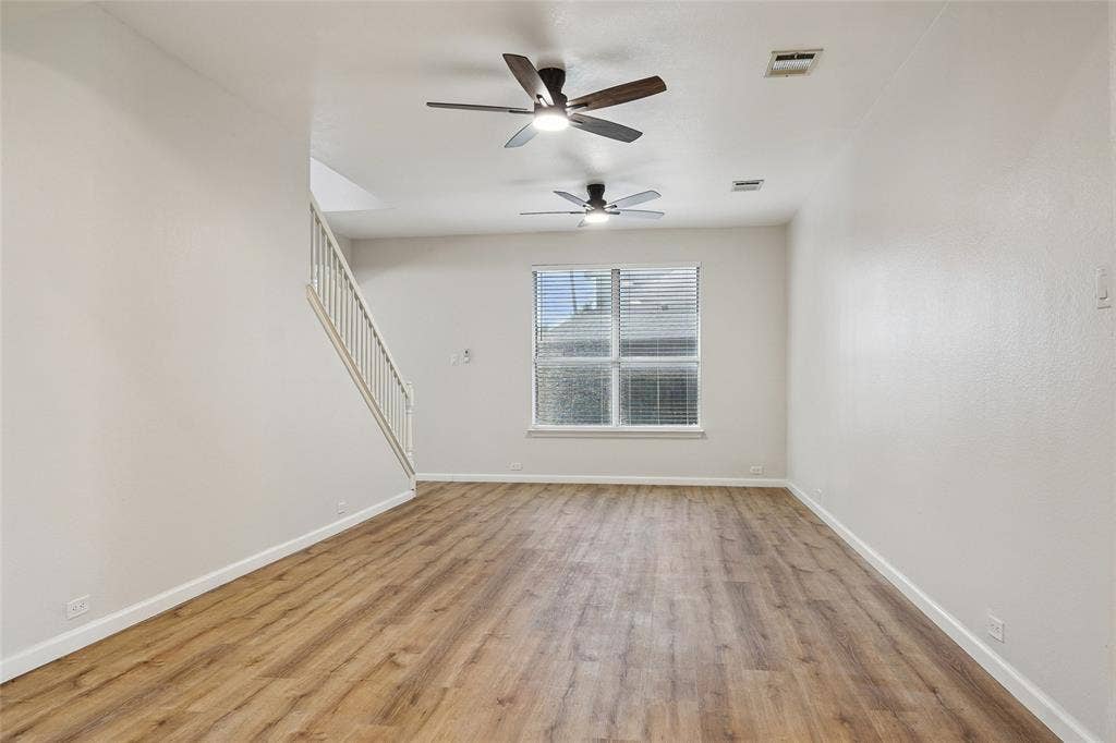 Wood-finish flooring, white walls, two ceiling fans with lighting, window with blinds, and staircase with white railing