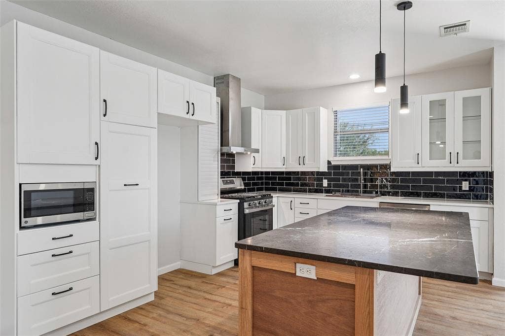 Contemporary kitchen featuring white cabinetry, a dark stone island countertop, a black subway tile backsplash, stainless steel appliances, and wood-finish flooring