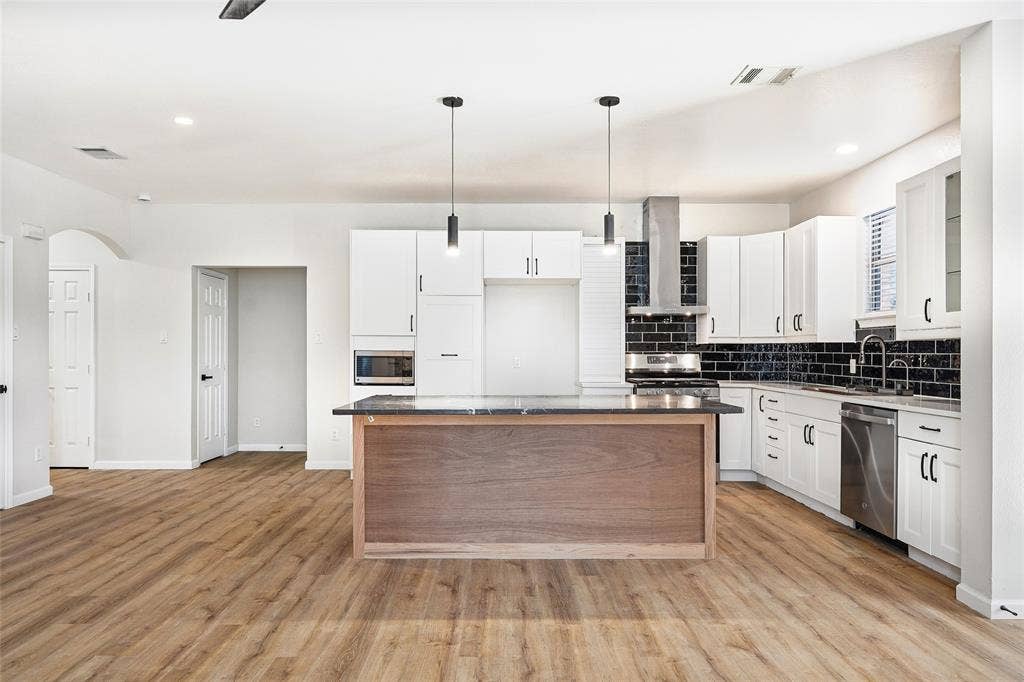 Kitchen featuring wood-finish flooring, white cabinetry, a dark tile backsplash, stainless steel appliances, and an island with a dark countertop and two pendant lights