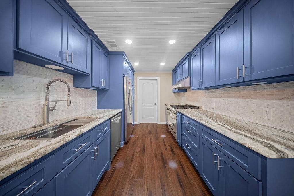 Galley kitchen featuring blue shaker-style cabinetry with brushed nickel hardware, light-toned stone countertops, and a full tile backsplash