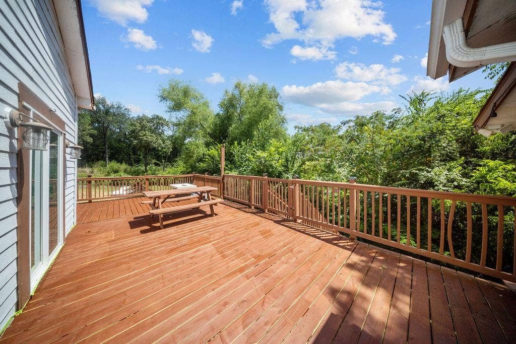 Spacious wood deck with natural wood railing, featuring a sliding glass door entry to the home's light blue siding, surrounded by mature trees
