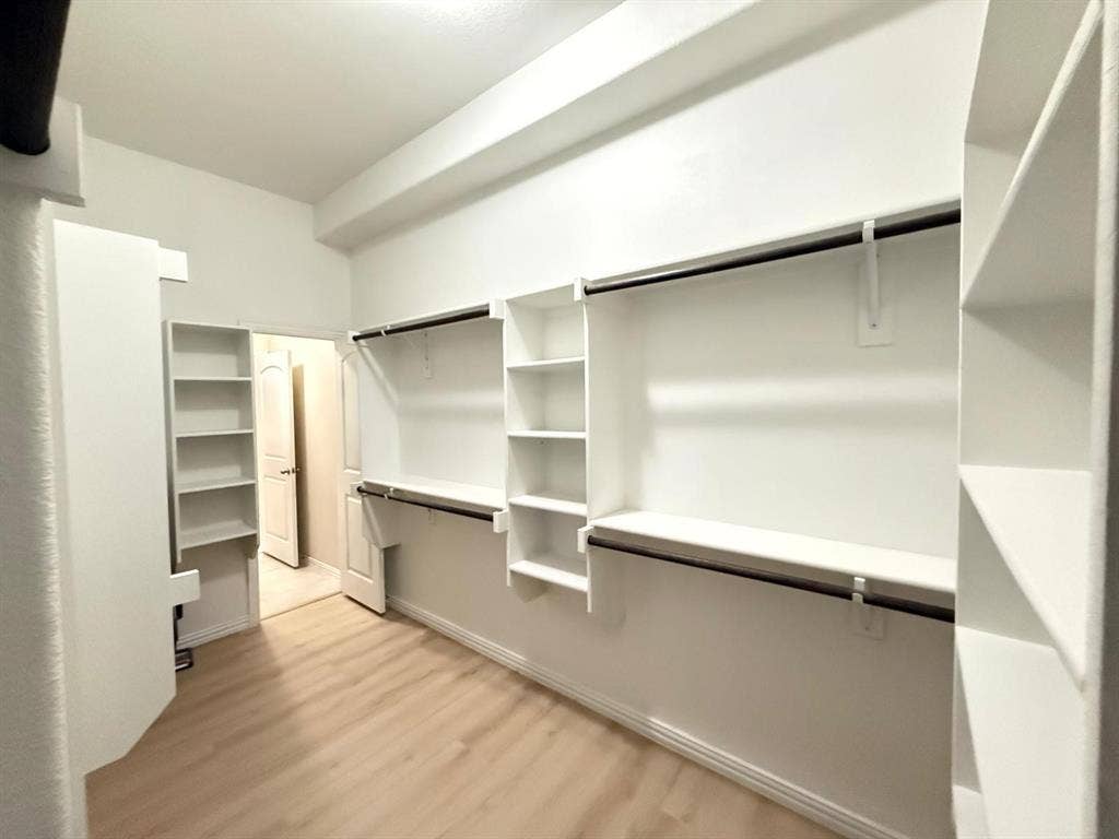 Spacious walk-in closet featuring light wood-finish flooring, white built-in shelving, and multiple hanging rods