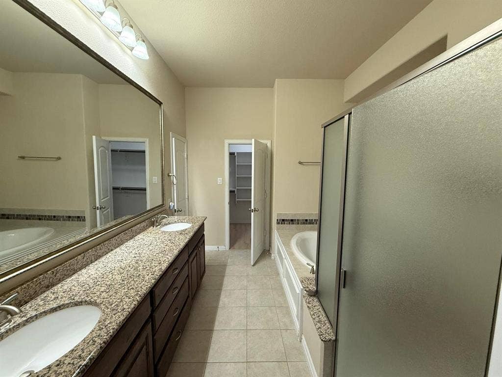 Bathroom featuring a long dual vanity with stone countertops, a full-width mirror, and an adjacent soaking tub with tile surround