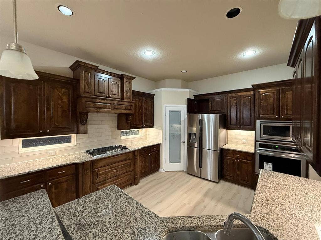 Spacious kitchen featuring dark wood cabinetry, granite countertops, stainless steel appliances, a gas range with a built-in hood, and light wood-finish flooring