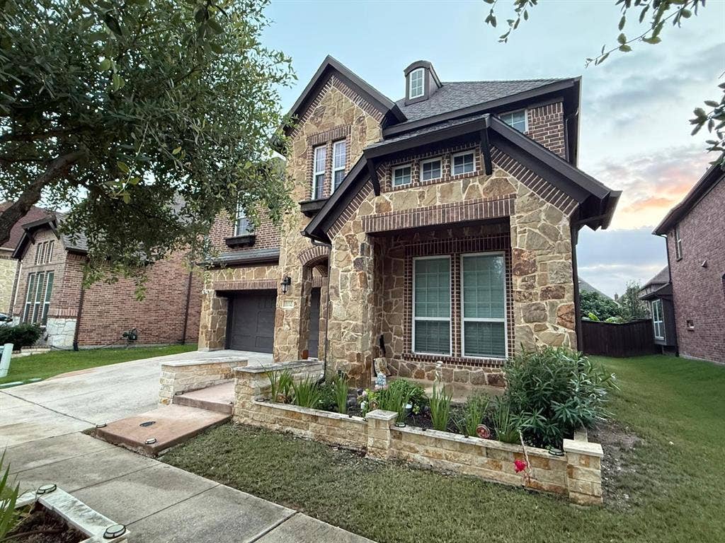 Two-story residence featuring a stone and brick facade, an attached garage, a dormer window, and a planted stone planter box
