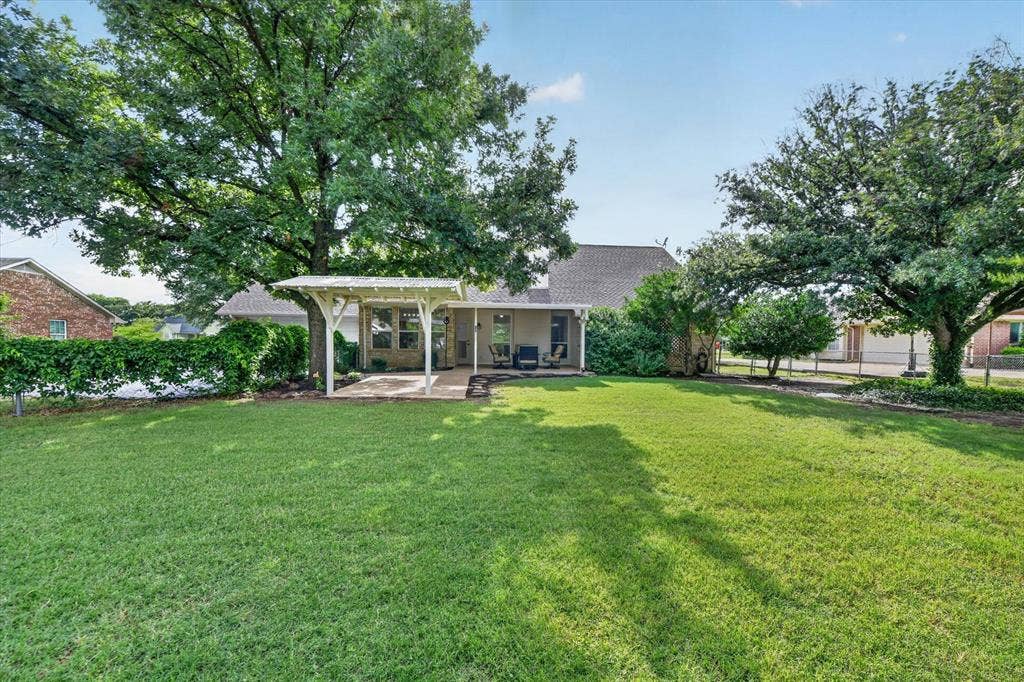 Expansive lawn with mature trees and a privacy hedge, featuring a covered patio with a white pergola structure and a concrete slab