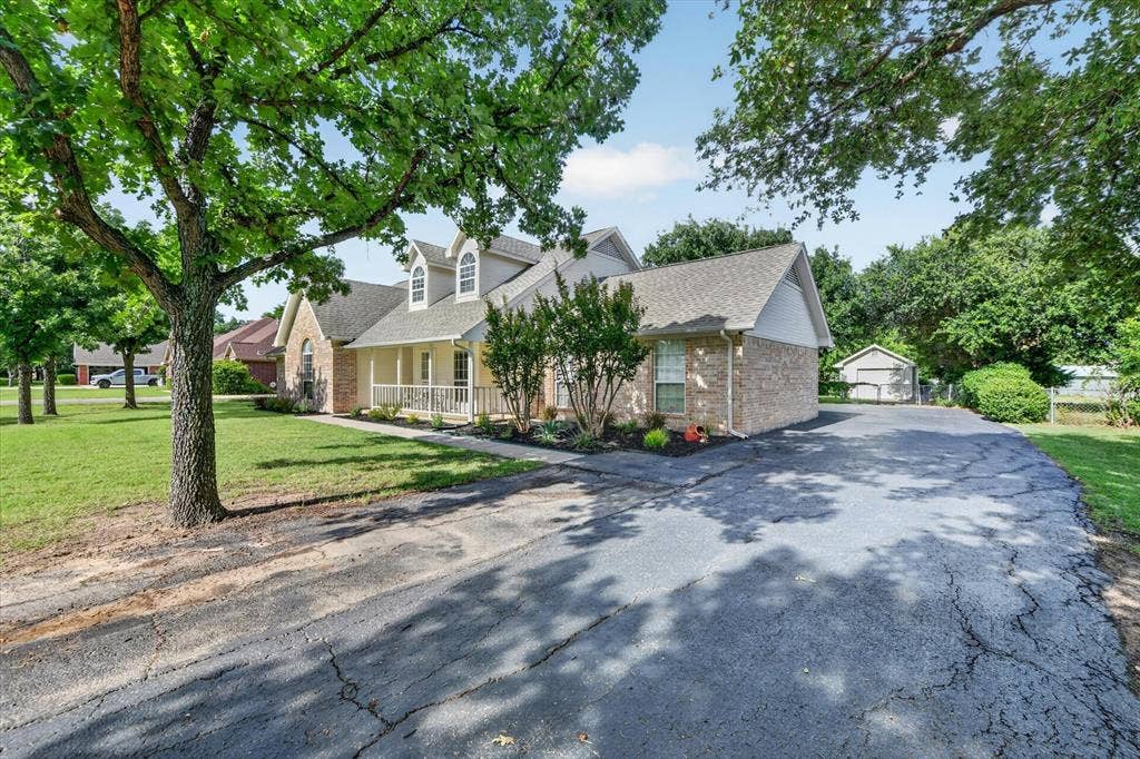 Traditional-style home featuring a brick facade, dormer windows, a covered front porch with white railings, mature trees, and an asphalt driveway leading to a detached shed