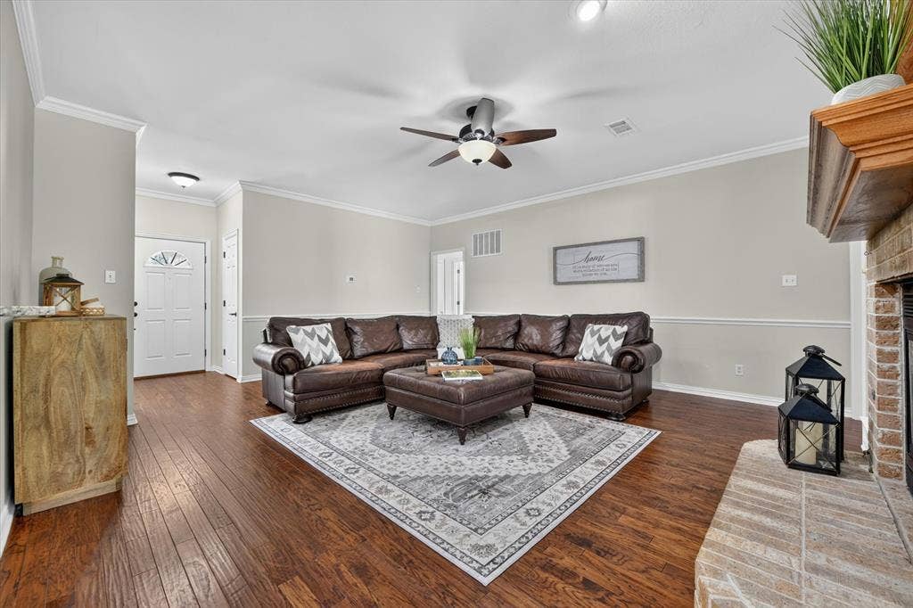 Spacious living area featuring wood-finish flooring, a brick fireplace with a wood mantel, crown molding, and a ceiling fan with integrated lighting