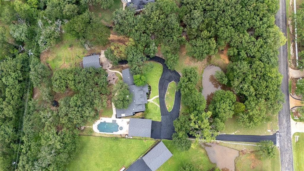 Aerial view showcasing a residential property with established trees, a main residence with a dark-toned roof, a detached structure with a matching roof, a swimming pool with surrounding patio space, and a curved asphalt driveway
