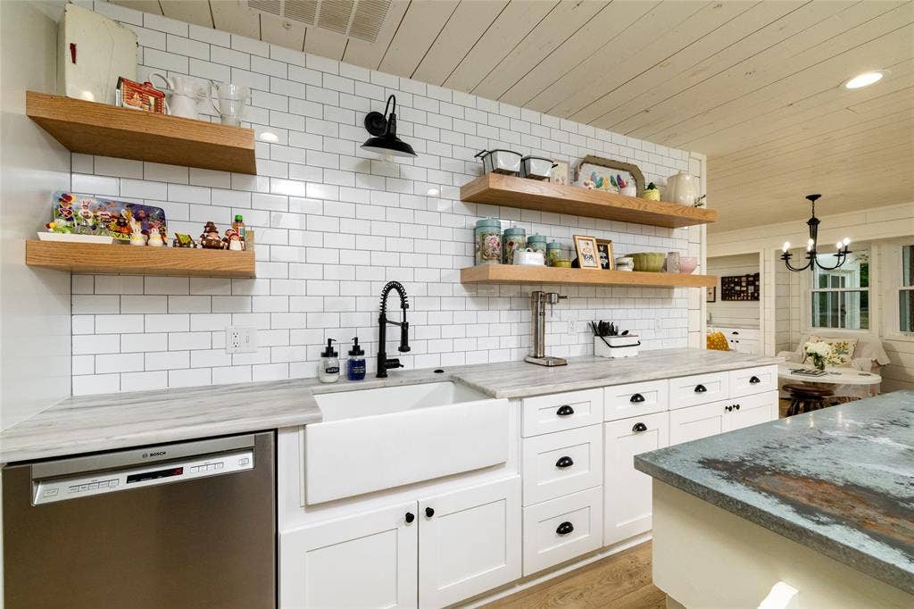Kitchen featuring white subway tile backsplash, white shaker cabinetry, an apron-front sink with a matte black faucet, wood-finish open shelving, and light countertops