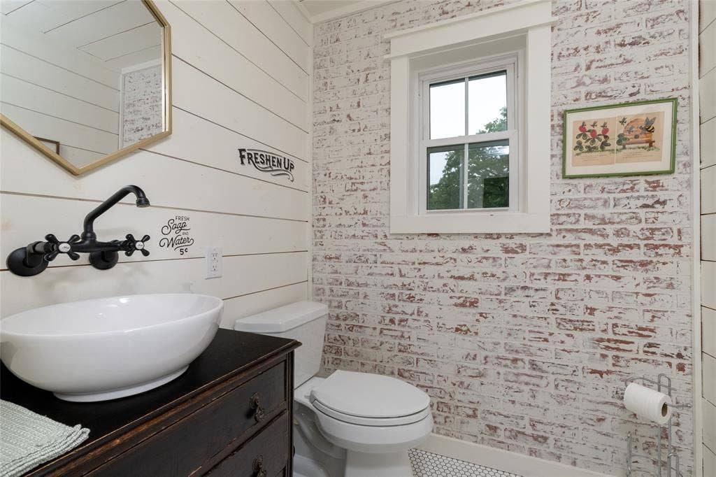Powder room featuring a vessel sink with a wall-mounted faucet, an antique-style vanity, ship-lap wall paneling, and a white-washed brick accent wall