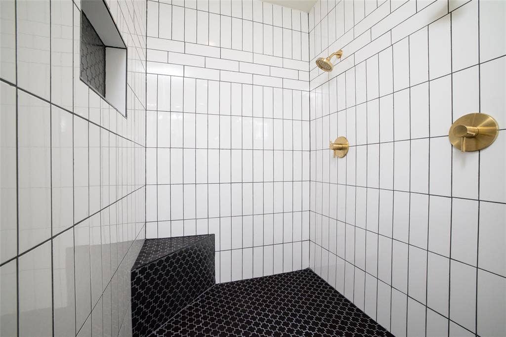 Tiled shower featuring vertical white subway tiles, a built-in bench with black patterned tiles, black patterned shower floor tiles, a recessed tile niche, and brass fixtures