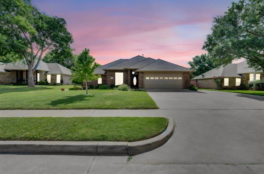 Brick exterior home featuring a two-car garage, concrete driveway, and front lawn with mature trees and landscaping
