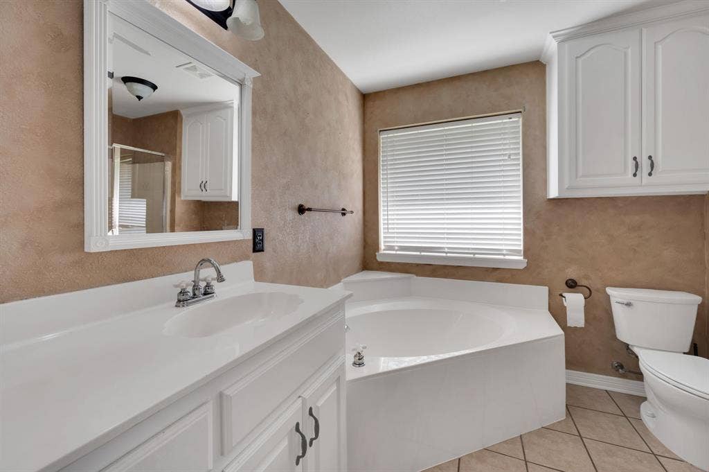 Bathroom featuring a built-in corner soaking tub, a white vanity with an integrated sink, a framed mirror, and upper white cabinetry