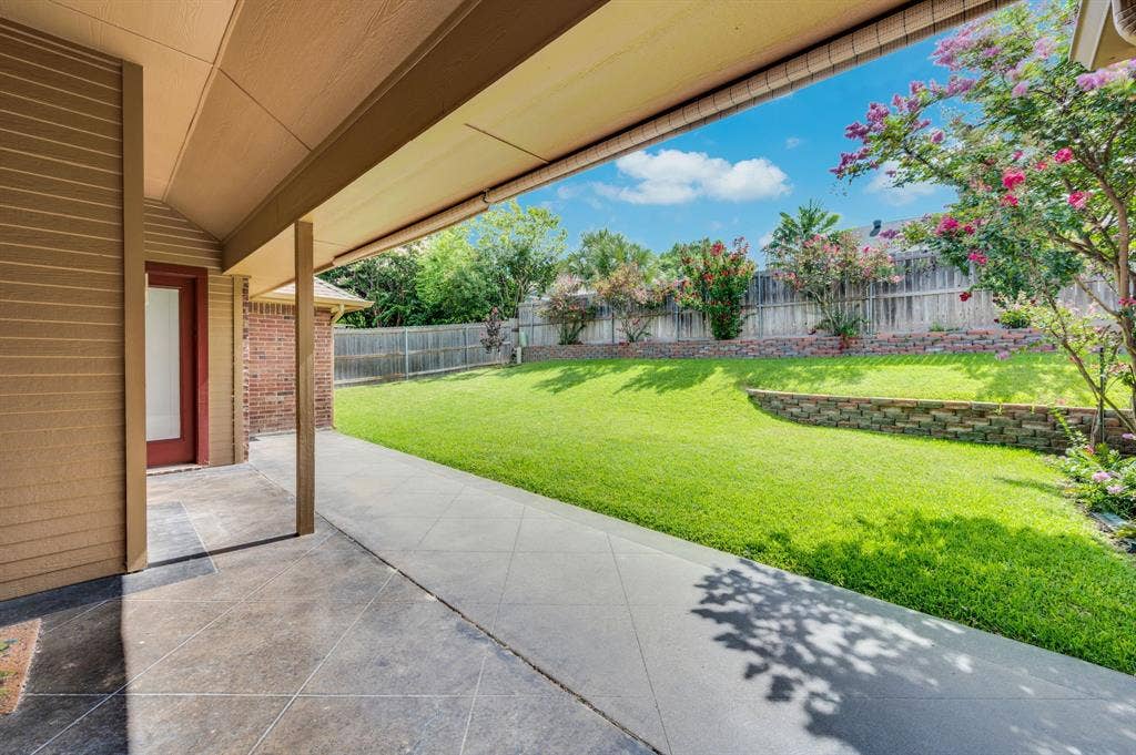 Covered concrete patio extending to a landscaped backyard with lush green lawn, a stone retaining wall, and mature flowering trees