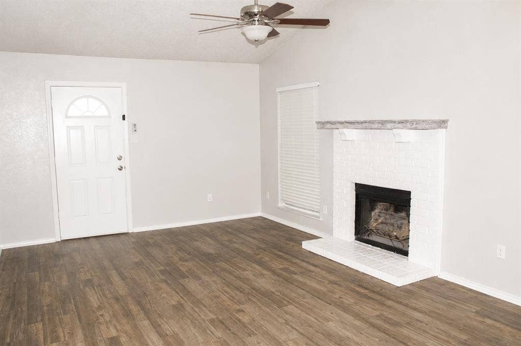 Spacious interior featuring wood-finish flooring, a white brick fireplace with mantel, white paneled entry door with arched transom, and a ceiling fan