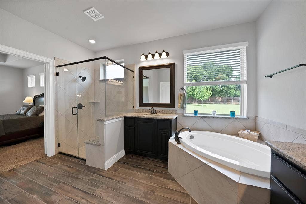 Spacious bathroom featuring wood-finish flooring, a frameless glass shower enclosure with diamond-patterned tile, a built-in soaking tub, a dark wood vanity with a stone countertop, and a window overlooking a green yard