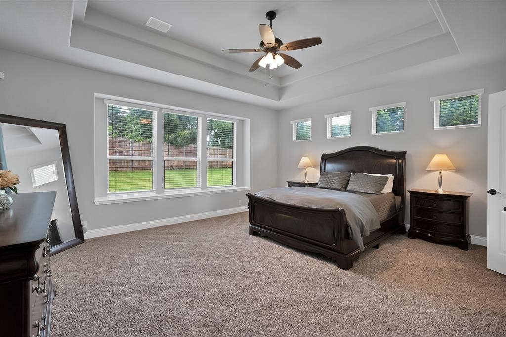 Spacious bedroom featuring a tray ceiling with a ceiling fan, plush carpet flooring, and a bank of three large windows providing ample natural light