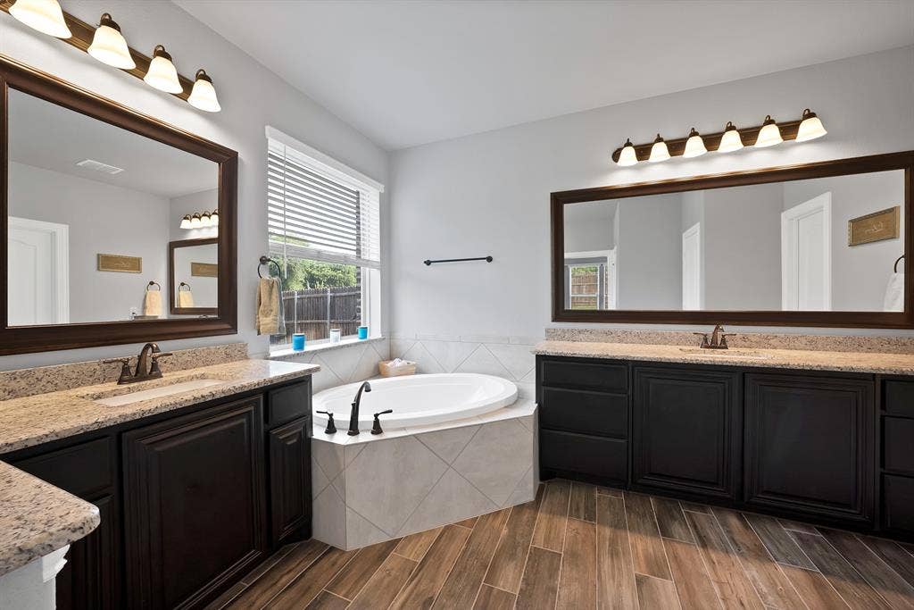 Spacious bathroom featuring a corner soaking tub, dual vanities with light-toned countertops, wood-finish flooring, and a large window with blinds