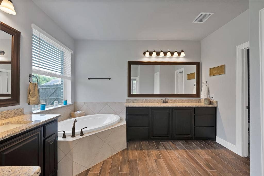 Soaking tub with tiled surround, dual vanities featuring granite countertops and dark wood cabinetry, wood-finish flooring, and a large window with blinds