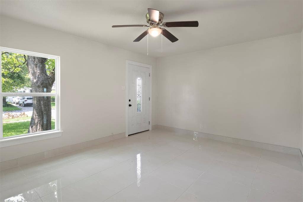 Spacious room featuring light-toned flooring, a ceiling fan with integrated lighting, a white entry door with decorative glass, and a large window providing outdoor illumination