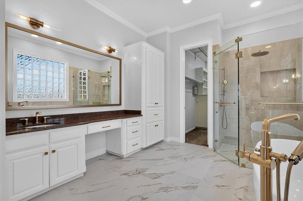 Spacious bathroom featuring a large vanity with white cabinetry and a brown countertop, a full-width mirror with brass lighting, a glass-enclosed shower with a rainfall showerhead, and a standalone tub with brass fixtures