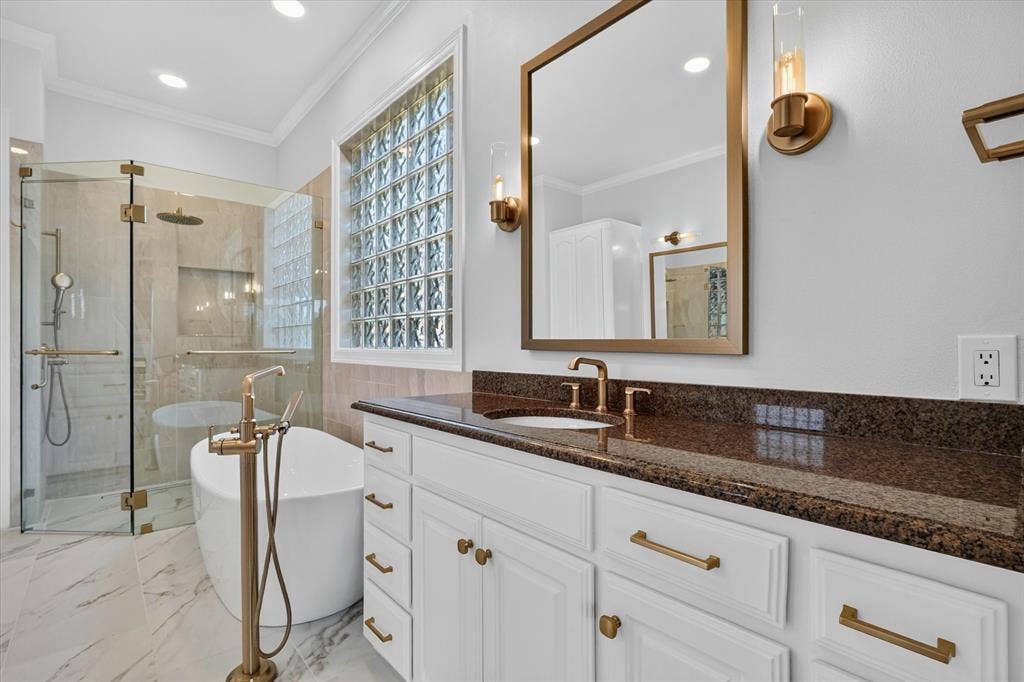 Bathroom featuring a freestanding tub with a brass floor-mounted faucet, a walk-in shower with glass enclosure and rain showerhead, a dual-sink vanity with a dark countertop and white cabinetry, a glass block window, and large format marble-look floor tiling