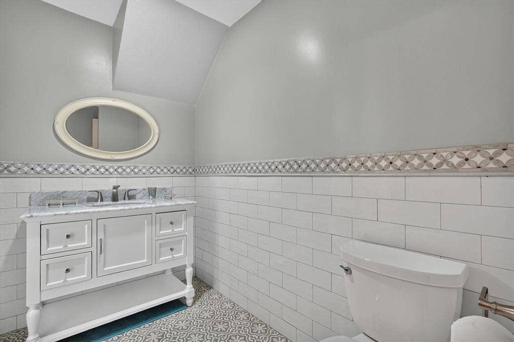 Bathroom featuring a white vanity with a stone countertop, a wall-mounted oval mirror, white subway tile wainscoting, decorative accent tile, and patterned floor tile
