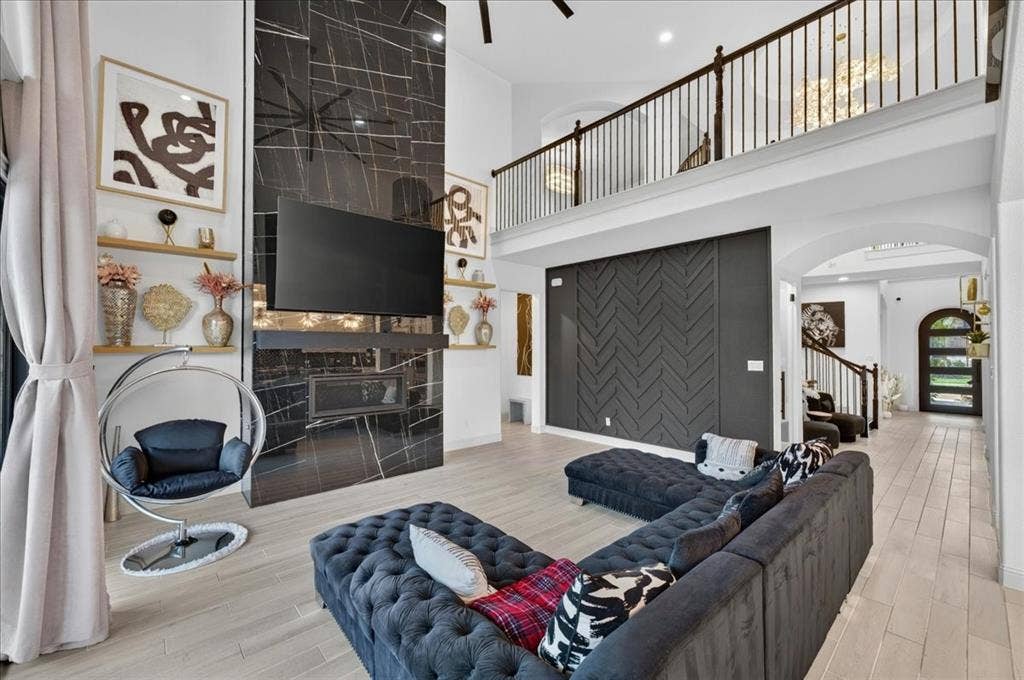 Two-story living area featuring a dramatic black stone-finish fireplace, light wood-finish flooring, a contemporary accent wall with chevron paneling, and an upper-level balcony with black iron railings