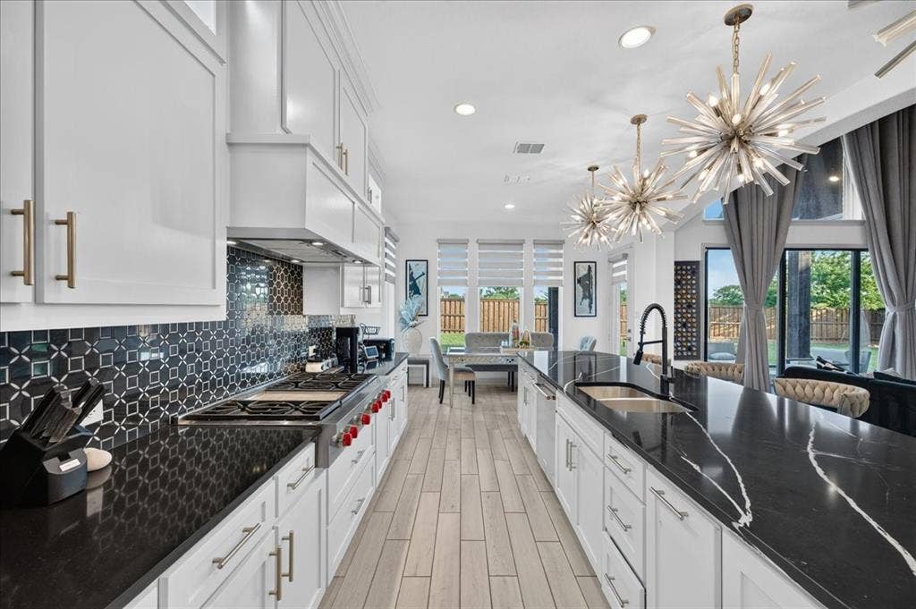 Spacious kitchen featuring an elongated island with integrated sink, black countertops with white veining, white cabinetry, a patterned black tile backsplash, and light wood-finish flooring
