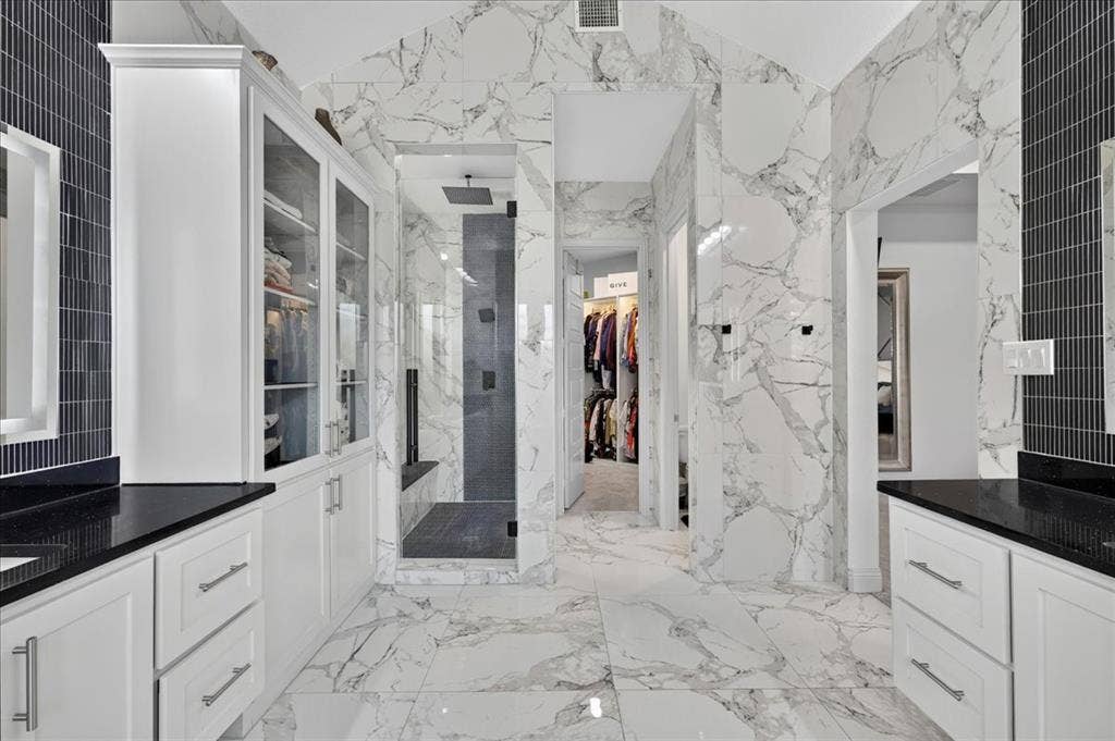 Spacious bathroom featuring white cabinetry with dark countertops, a walk-in shower with a glass enclosure, white marble-look tile flooring, and white marble-look tile walls