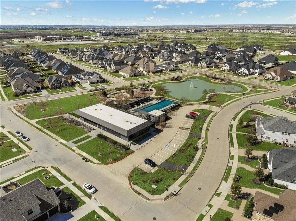Aerial view of a residential community featuring a modern amenity building, an outdoor swimming pool, and a central pond with a fountain