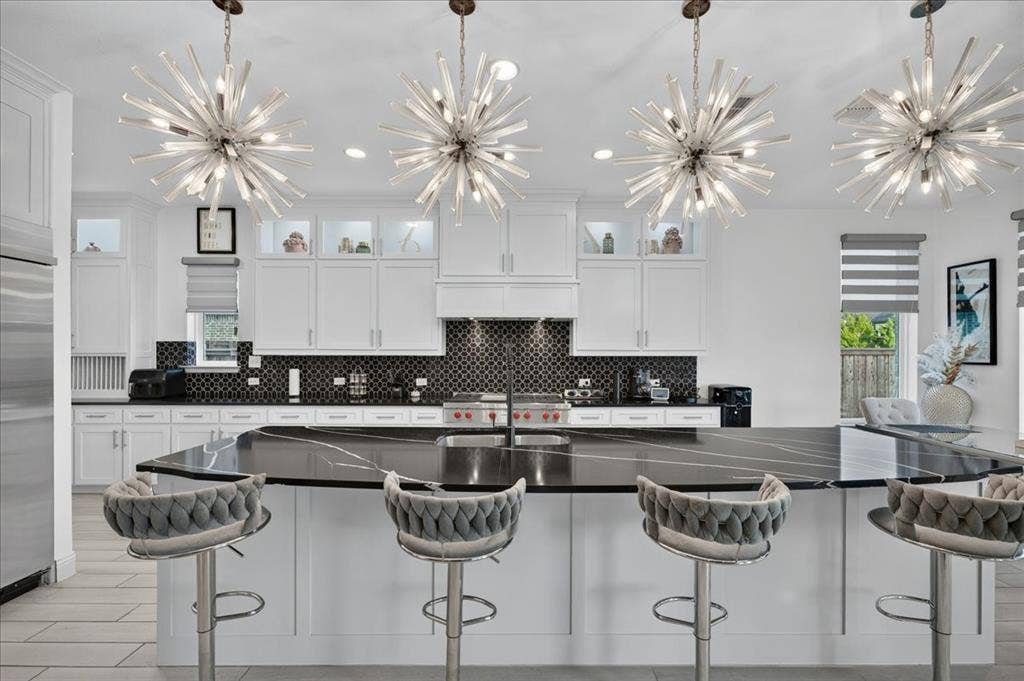 Gourmet kitchen featuring a large central island with a dark countertop, white perimeter cabinetry, a mosaic tile backsplash, and four contemporary chandeliers