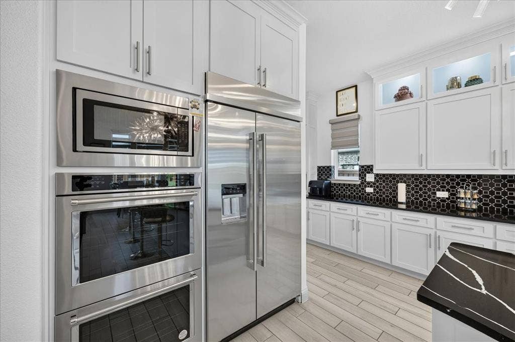 Gourmet kitchen featuring stainless steel appliances, a built-in oven and microwave, light wood-finish flooring, white cabinetry, and a black patterned tile backsplash
