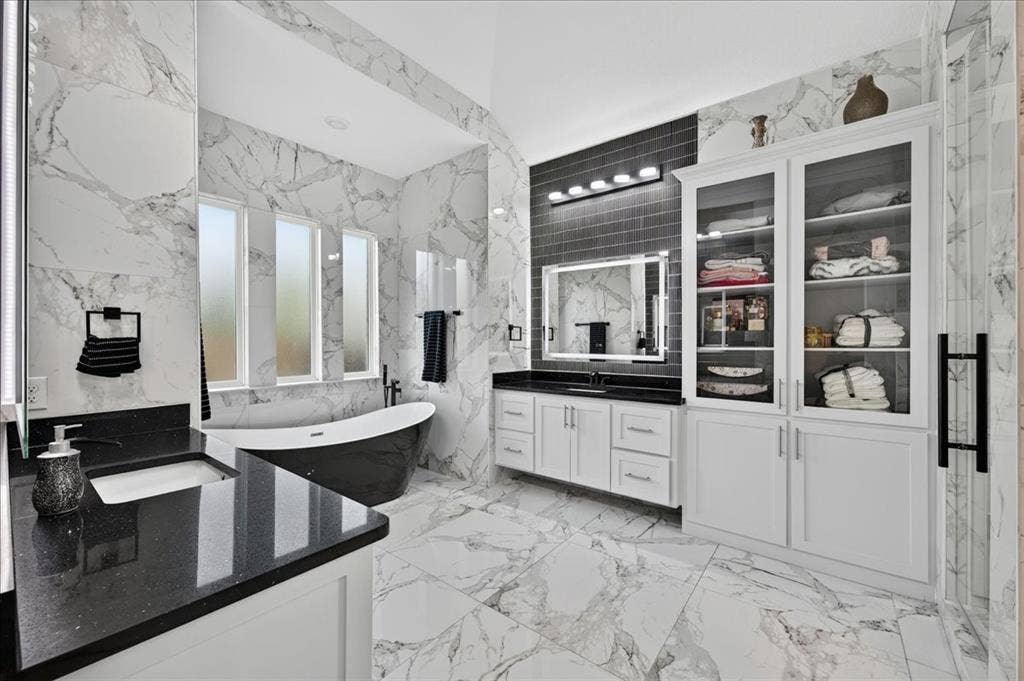 Spacious bathroom featuring marble-look tile flooring and walls, a freestanding black soaking tub, a dual vanity with dark countertops and white cabinetry, built-in storage cabinets with glass doors, and contemporary lighting fixtures
