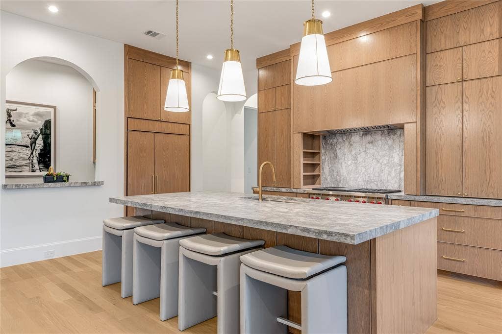 Center island with a stone countertop, three pendant lights, wood-finish flooring, full-height wood cabinetry, and an arched wall opening