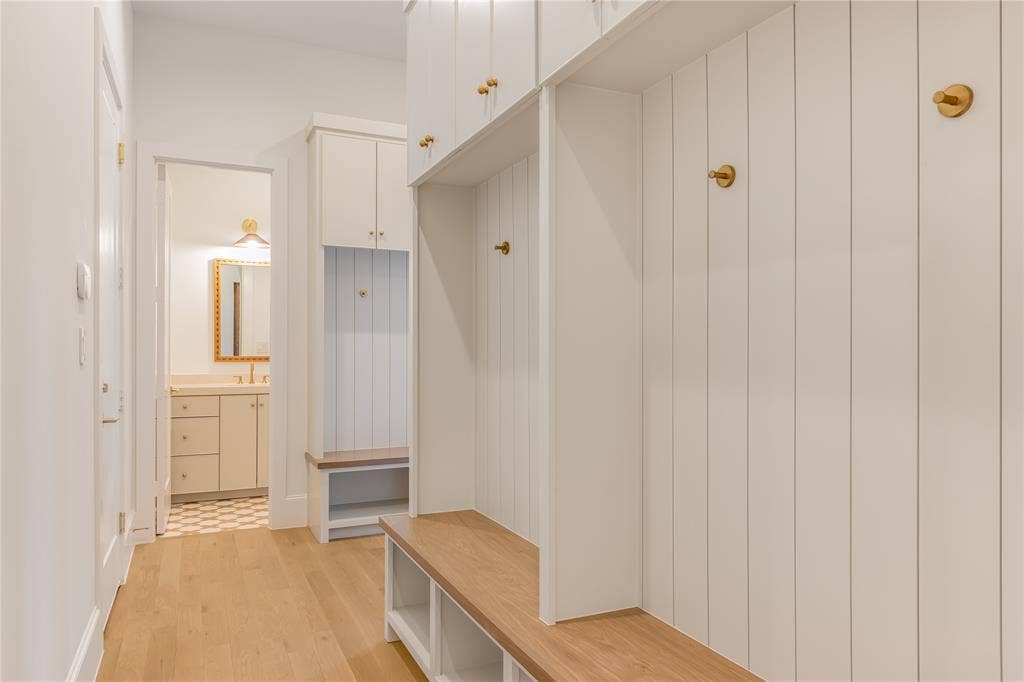 Built-in storage bench with wood-finish top and open shelving, vertical paneled wall accents, brass-finish hooks, overhead cabinetry, and light wood-finish flooring