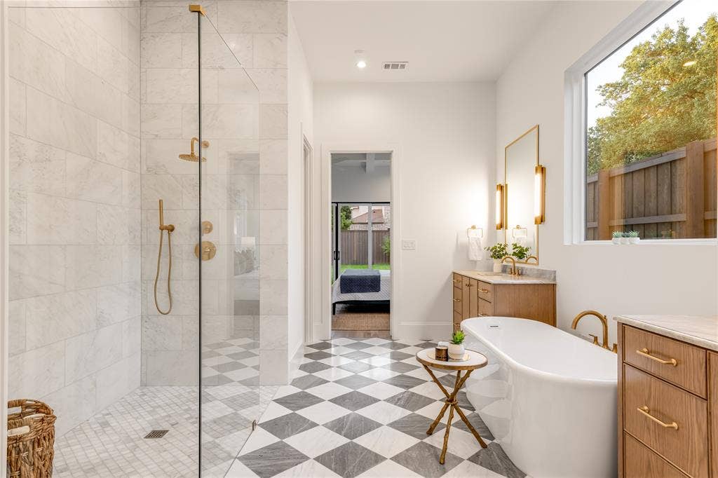 Glass-enclosed shower with gold-tone fixtures, freestanding soaking tub, wood-finish vanity with stone countertop, checkerboard tile flooring, and a large window