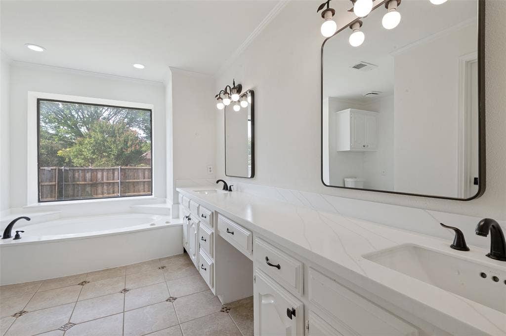 White double vanity with integrated sinks and black fixtures, featuring a large soaking tub with a surrounding window, tile flooring, and illuminated mirrors